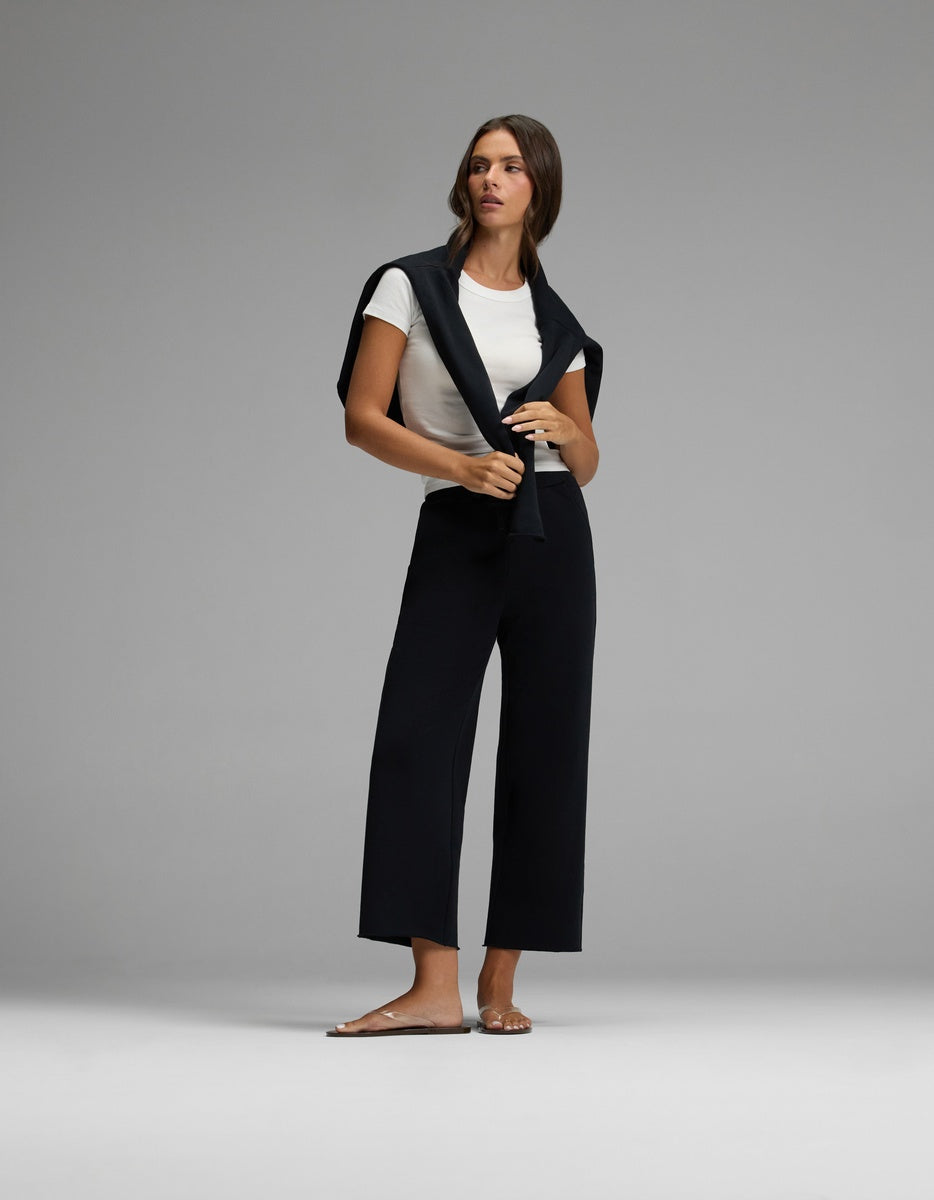 A woman stands barefoot against a plain gray background, wearing Cozy Earth’s Women’s Chelsea Cropped Pant in black, paired with a white T-shirt and a black sweater draped over her shoulders.