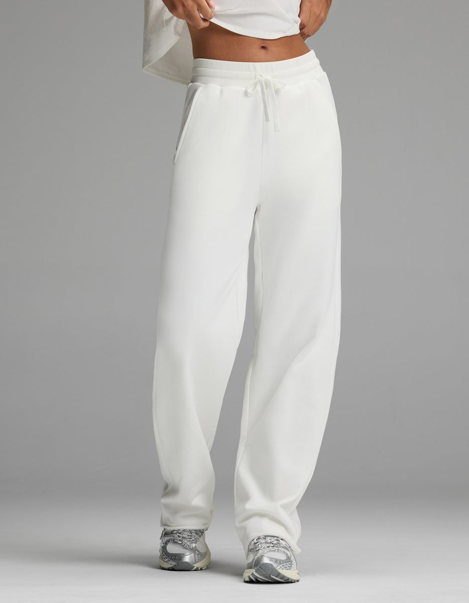 A person stands against a plain gray background wearing the Cozy Earth Women’s Chelsea Pant in Bone and a white cropped top, paired with gray sneakers. Their face is not visible.