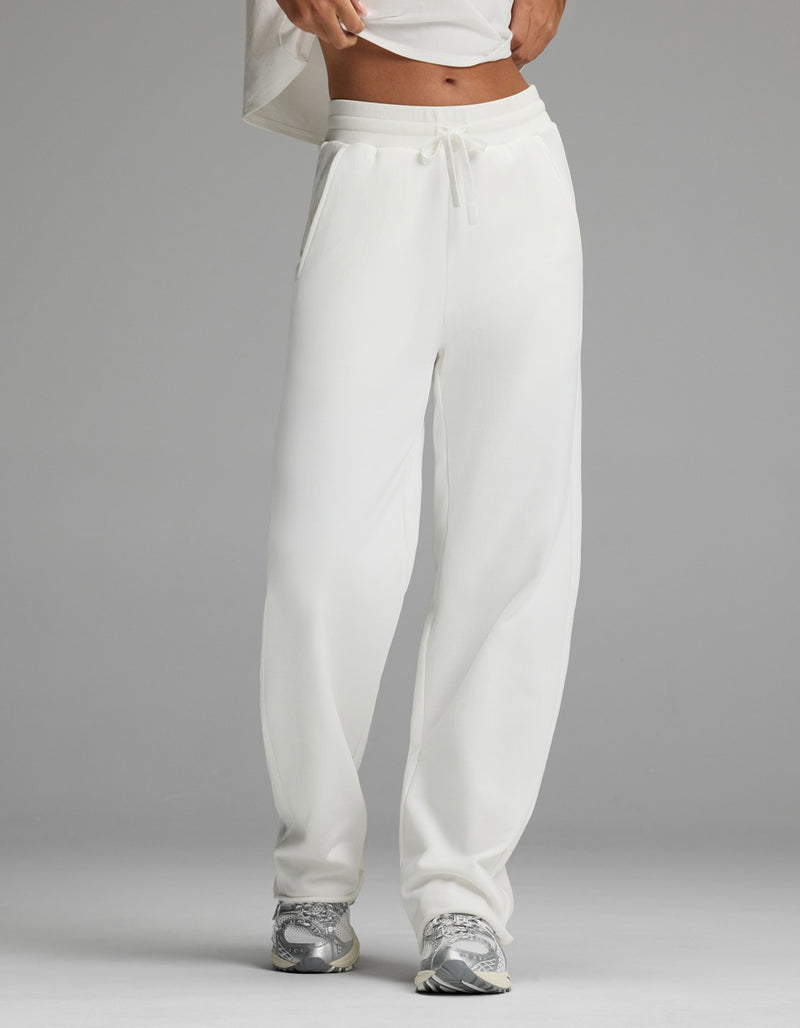 A person stands against a plain gray background wearing the Cozy Earth Women’s Chelsea Pant in Bone and a white cropped top, paired with gray sneakers. Their face is not visible.