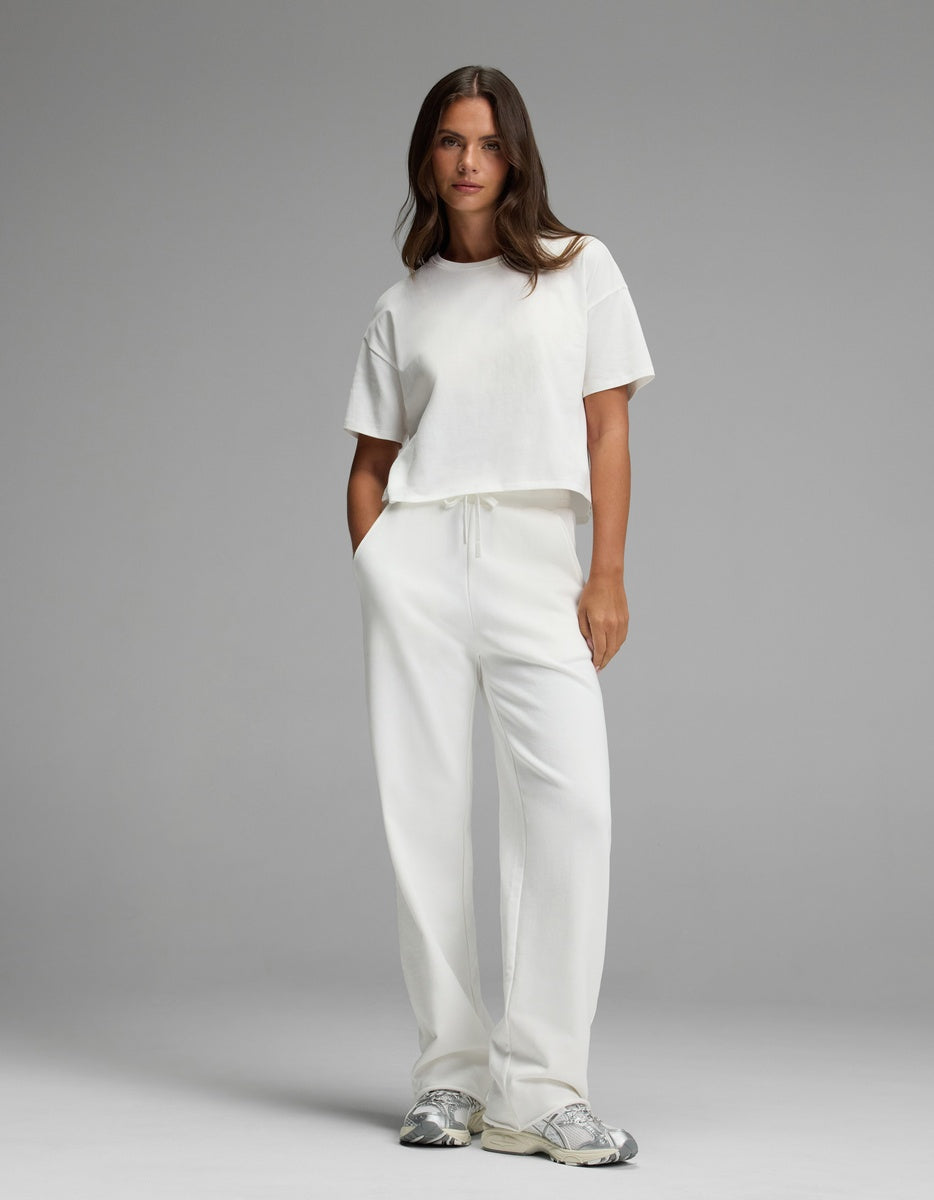 A woman stands against a gray background with her hands in her pockets and a neutral expression, wearing Cozy Earth’s Women’s Chelsea Pant in white paired with a white t-shirt and silver sneakers.