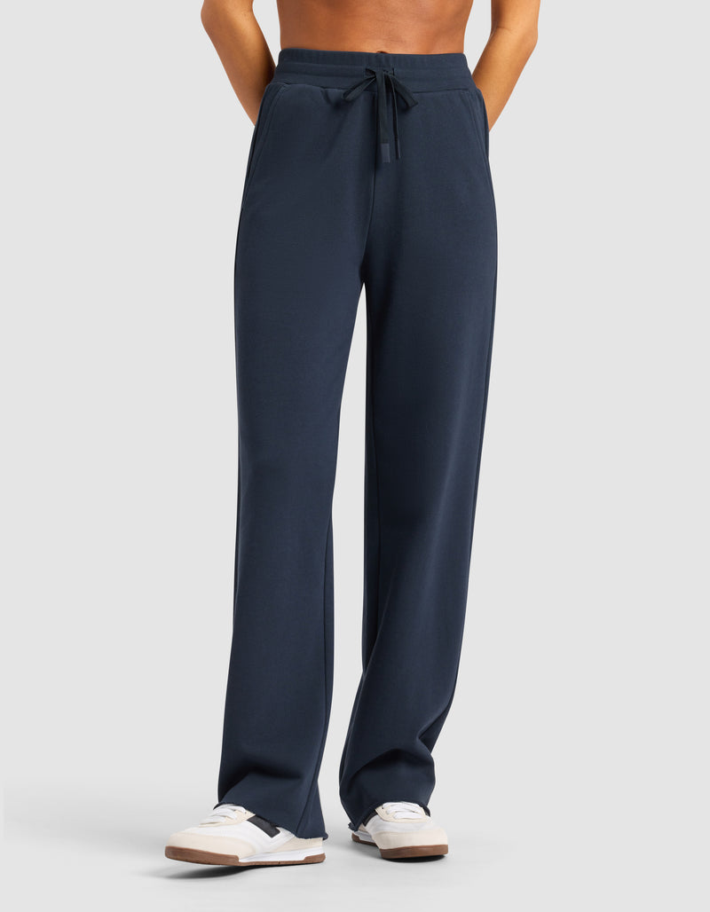 A person models the Cozy Earth Chelsea Pant in navy blue, paired with white sneakers, standing hands-in-pockets and facing forward against a plain light gray background.