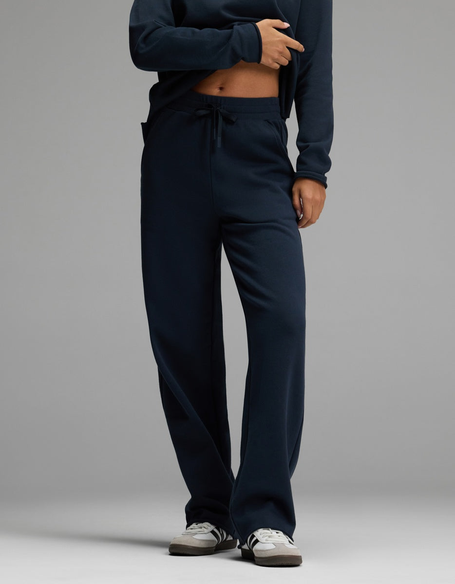 A person wearing Cozy Earth’s Women’s Chelsea Pant in Eclipse, paired with a matching sweatshirt and white sneakers, stands against a plain gray background. Their hand rests near the waistband of the pants, and their face is not visible in the image.