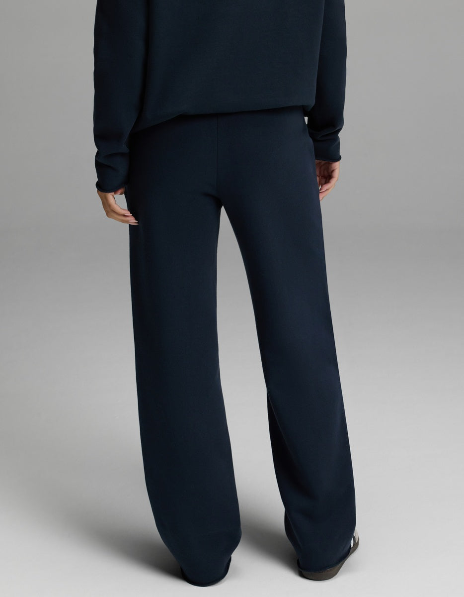 A person is pictured from behind, indoors against a plain light gray background, wearing Cozy Earth’s Women’s Chelsea Pant paired with a dark navy sweater. The lower half of the body is visible.