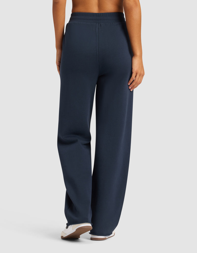 A person faces away from the camera in Cozy Earth's Chelsea Pant, featuring an elastic waistband and relaxed, straight-leg fit in dark navy. They pair it with white sneakers against a plain, light background.