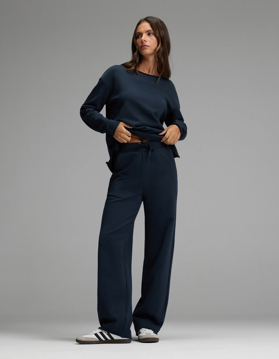 A woman stands against a plain background, wearing the Cozy Earth Women’s Chelsea Pant and a loose-fitting dark navy sweater, paired with black and white Adidas sneakers.