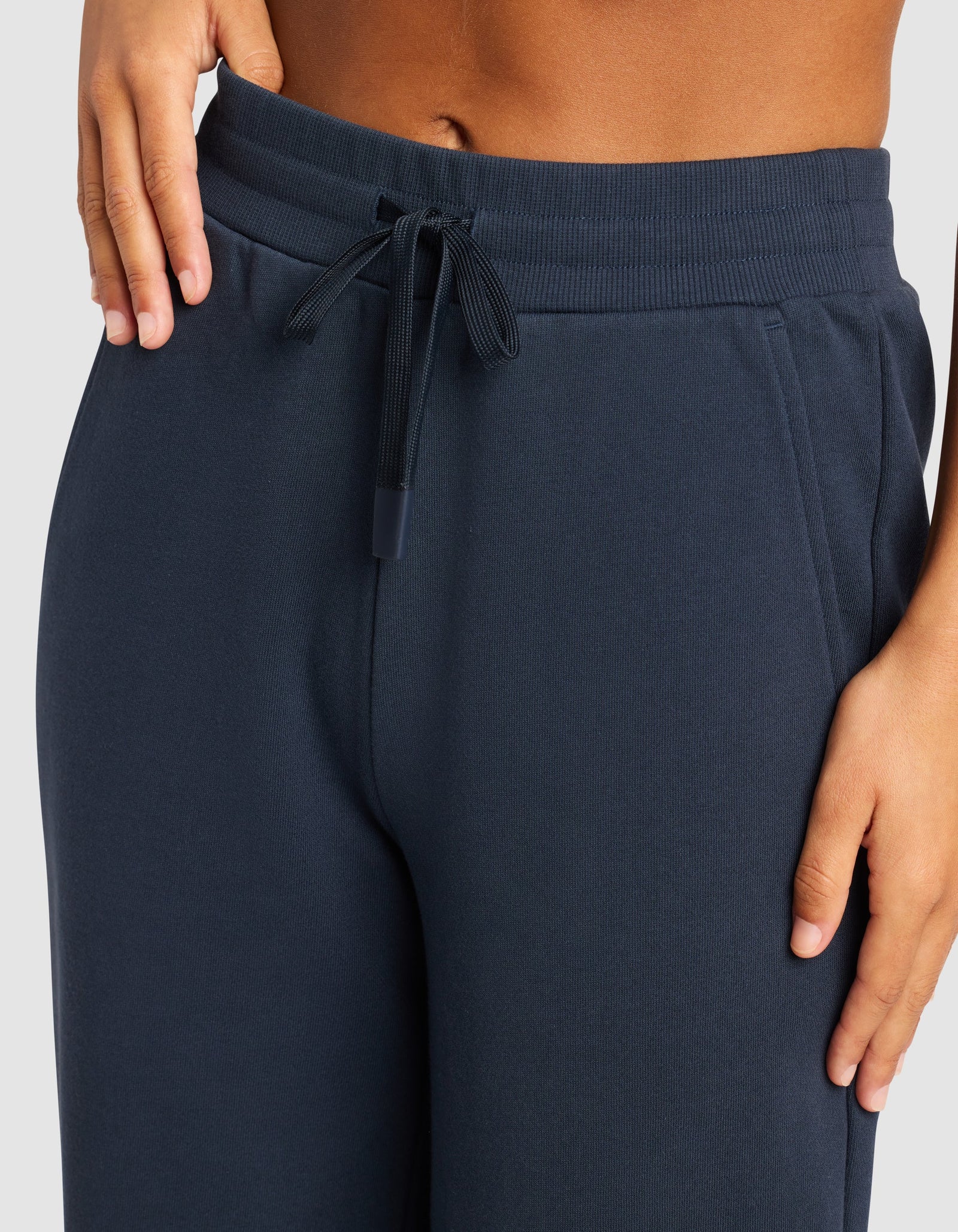 A close-up shows a person wearing Cozy Earth’s Chelsea Pant in dark blue with a drawstring waist, one hand on the waistband and the other at their side, against a plain light gray background.