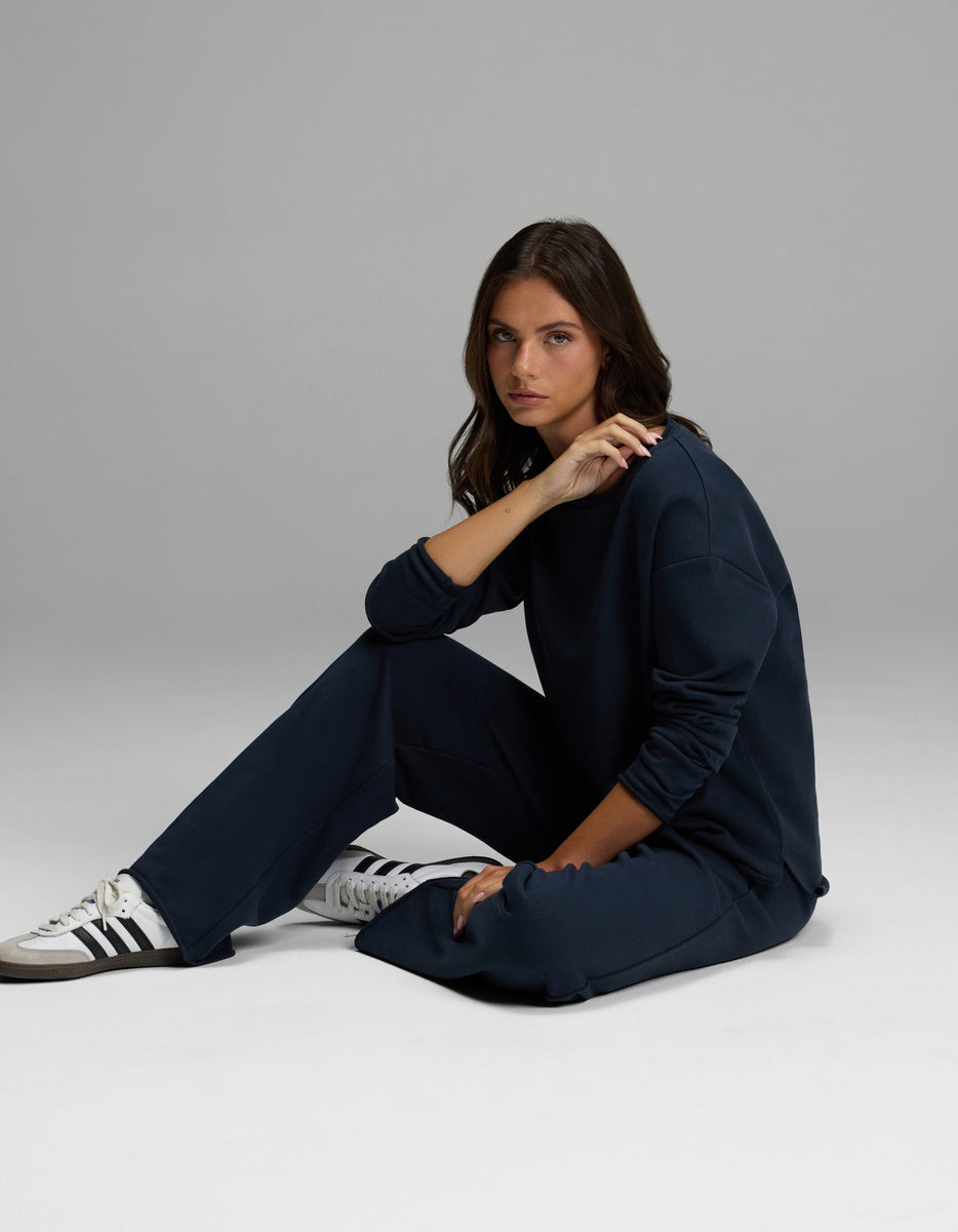 A person with long brown hair sits on the floor in a relaxed pose, wearing the Cozy Earth Women’s Chelsea Pant in navy blue, paired with a matching sweatshirt and white sneakers featuring black stripes. The background is plain and gray.