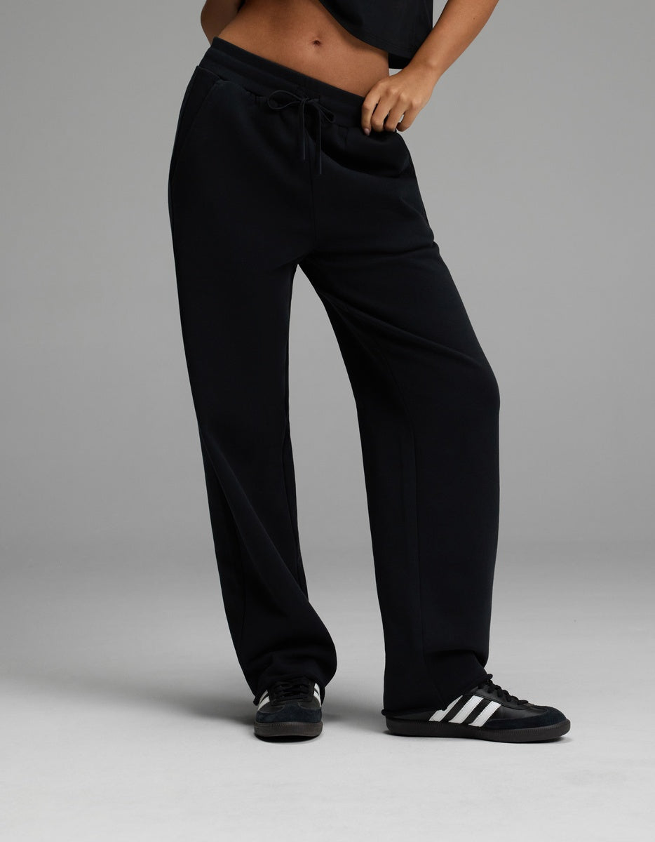 A person wearing the Cozy Earth Women’s Chelsea Pant in Jet Black pairs it with a black top and black sneakers with white stripes, standing with one hand in a pocket against a plain gray background.