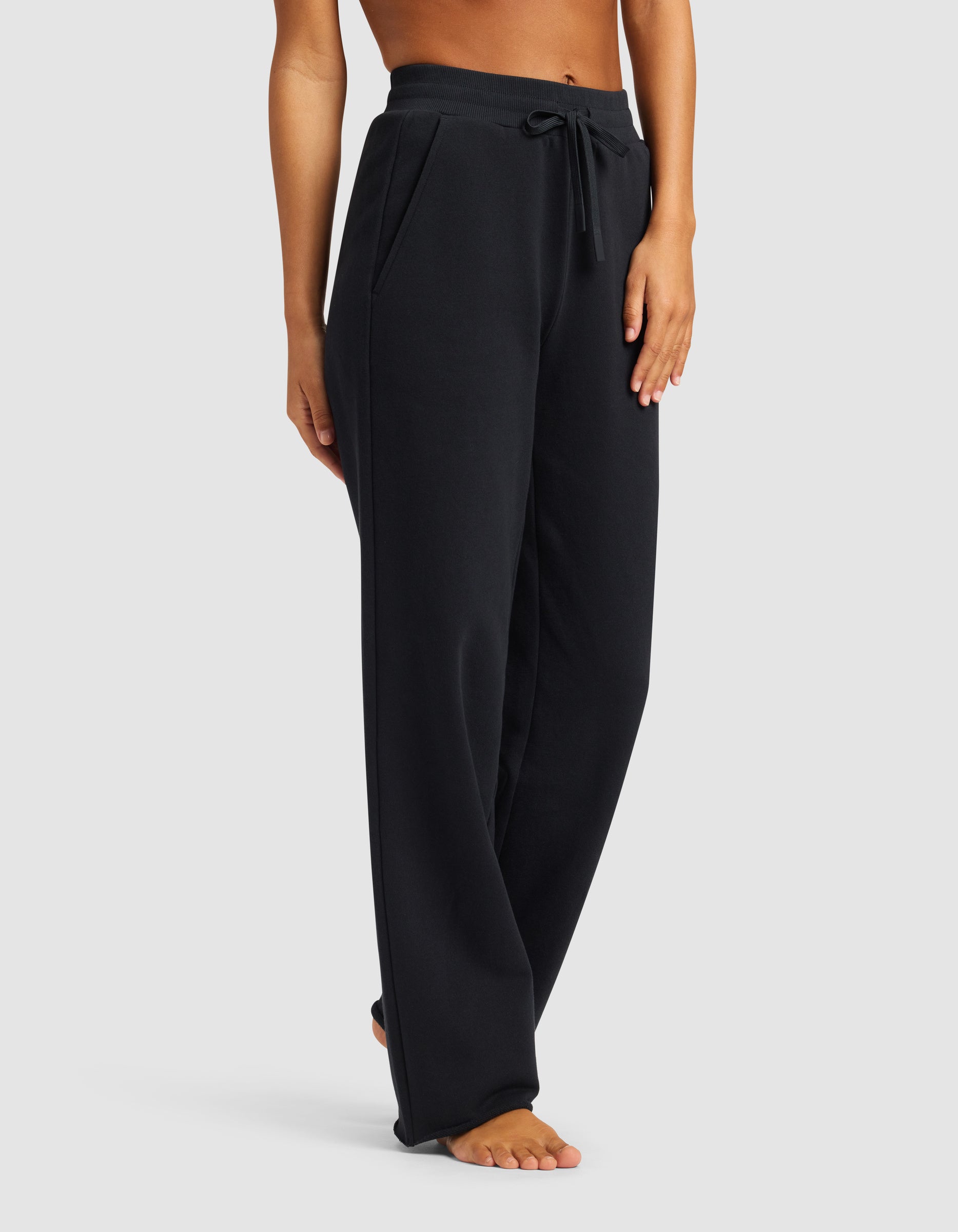 A person is seen barefoot against a plain light background, wearing Cozy Earth’s Chelsea Pant—loose-fitting black sweatpants with a drawstring waist. The upper body is cropped out of the frame.