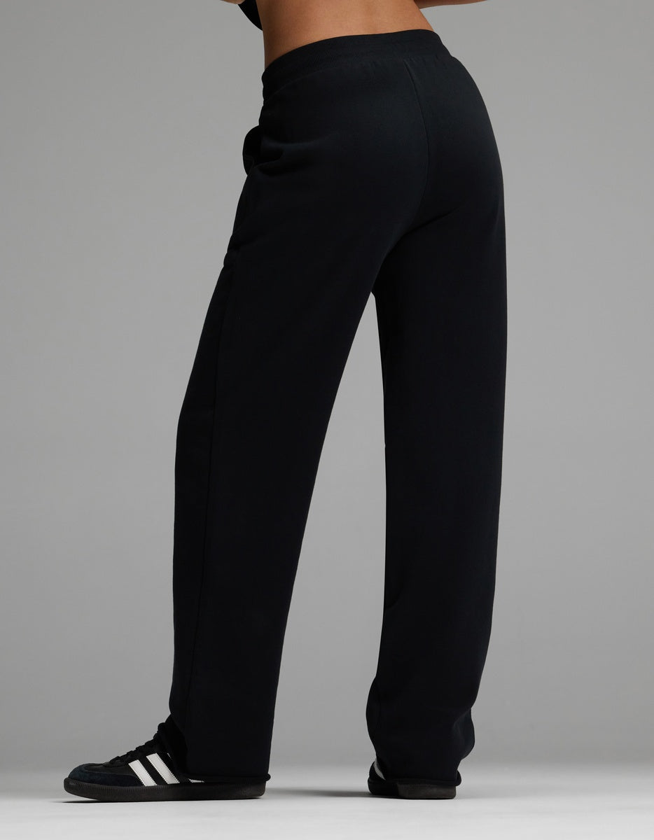 A woman is shown from behind against a gray background, wearing Cozy Earth’s Women’s Chelsea Pant in black and black Adidas sneakers with white stripes, with the image focused on the lower half of her body.