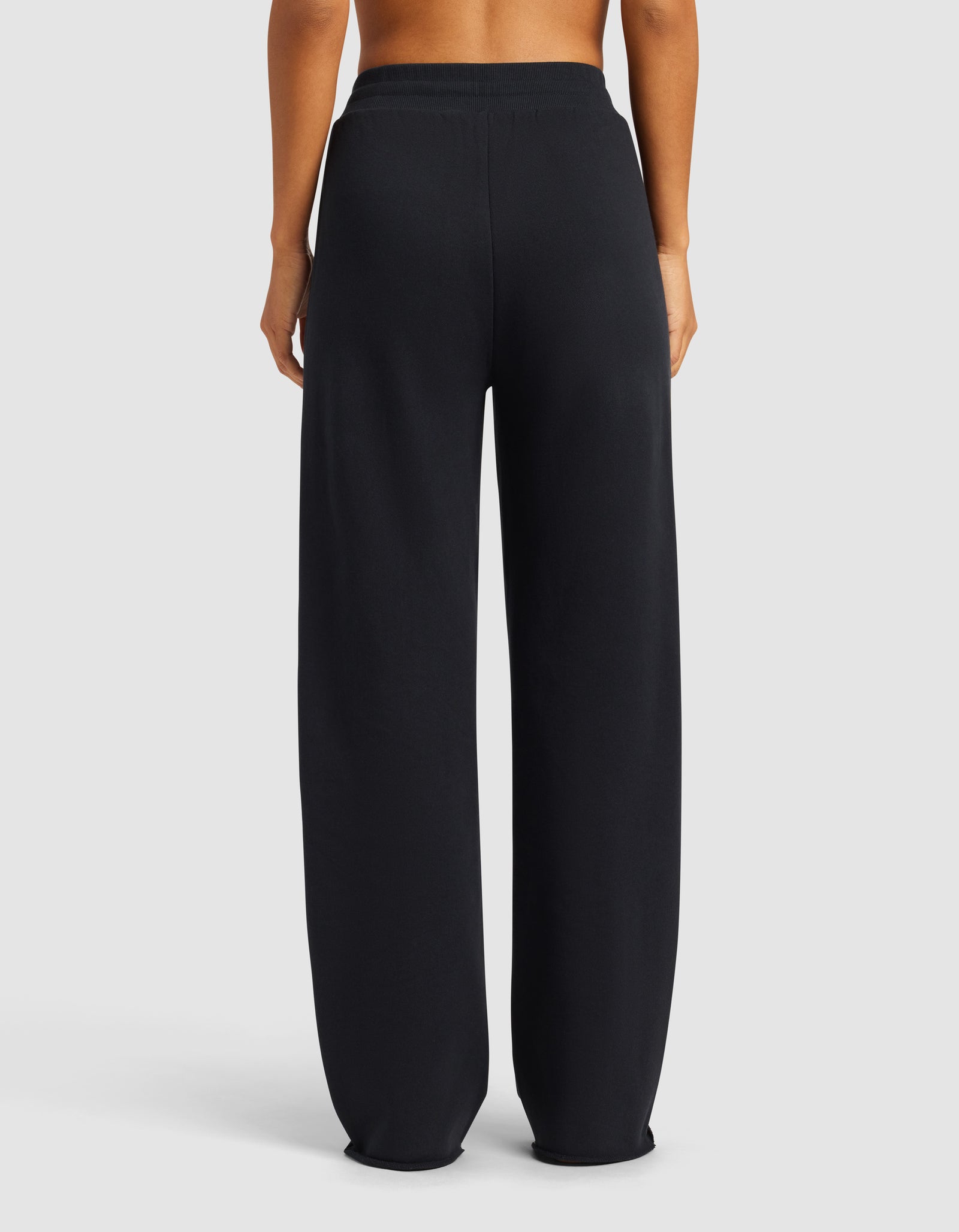 A person is shown from the waist down, standing and facing away, wearing Cozy Earth's Chelsea Pant in black with an elastic waistband. The background is a plain light gray.