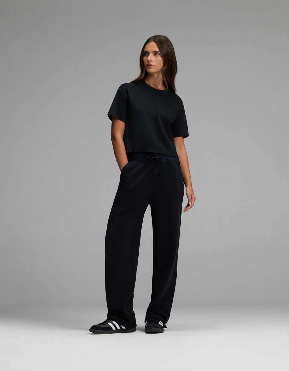 A woman stands against a gray background, wearing black Cozy Earth Women’s Chelsea Pants, a black t-shirt, and black Adidas sneakers with white stripes. She has her hands in her pockets and looks slightly to her left.