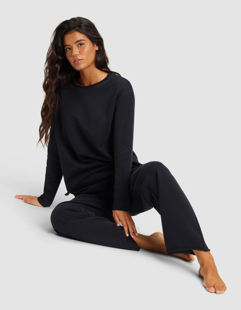 A woman with long dark hair wears a loose black long-sleeve top and Cozy Earth’s Chelsea Pant, sitting on the floor with one leg bent and the other extended against a plain light background.