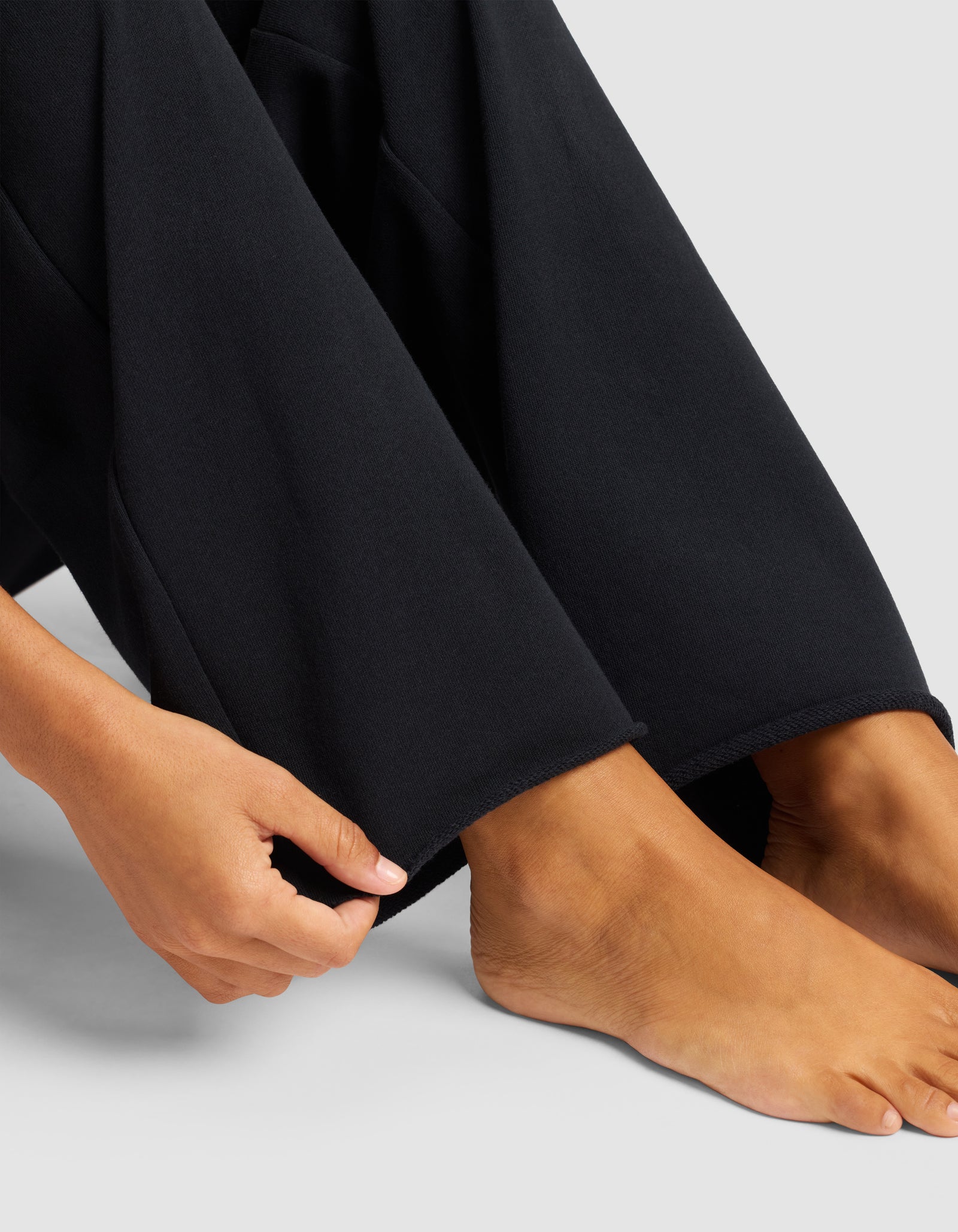 A person sits on the floor wearing Cozy Earth's Chelsea Pant in black. Their hands rest near their ankles, and their bare feet are visible against a light background.