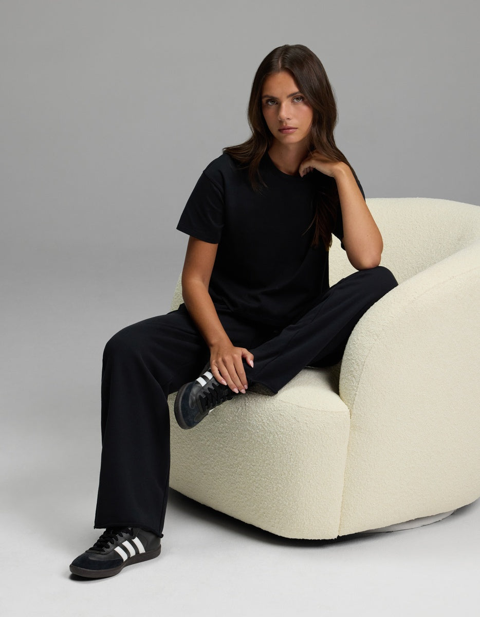 A woman with long brown hair, wearing a black t-shirt, black sneakers, and the Cozy Earth Women’s Chelsea Pant, sits on a cream textured armchair against a plain gray background.