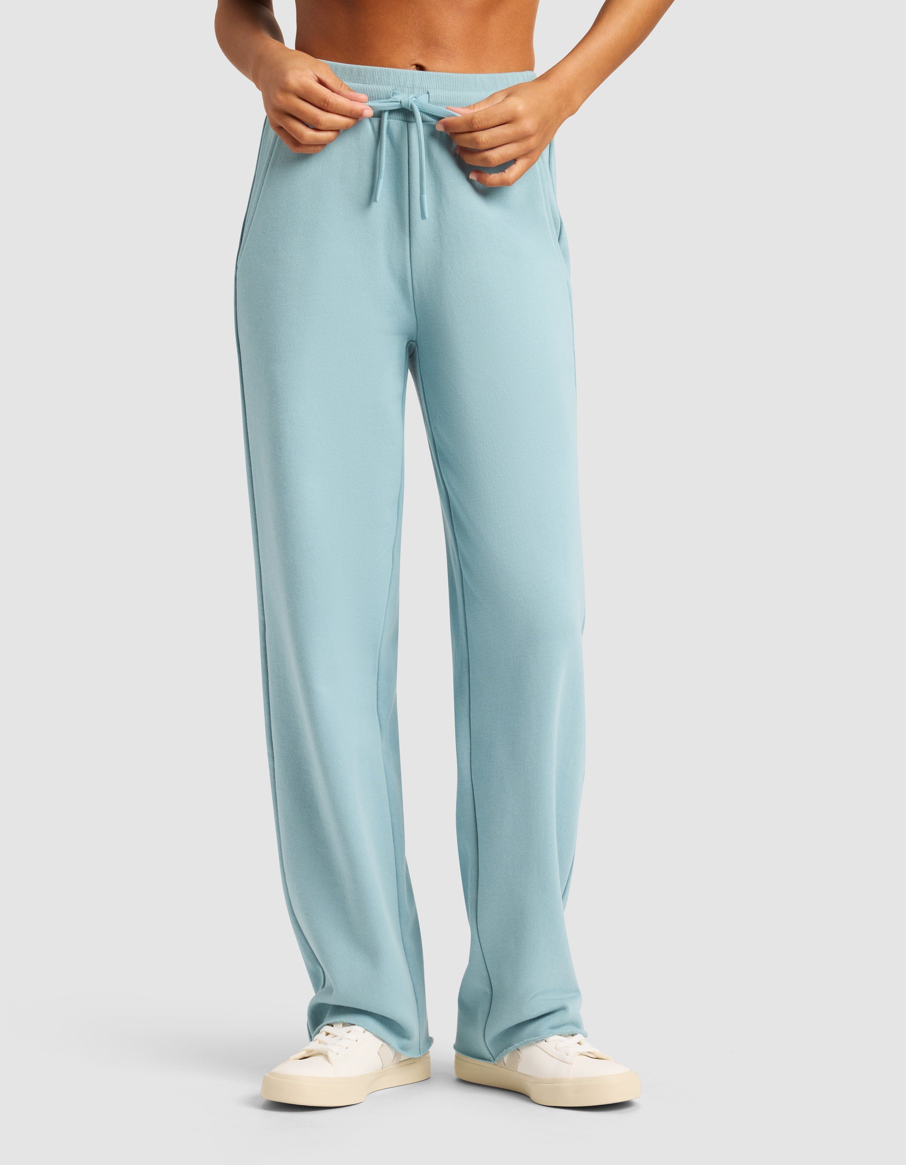 A person models the Cozy Earth Chelsea Pant in light blue, paired with white sneakers against a plain background, adjusting the waistband. Only their lower torso and legs are visible.