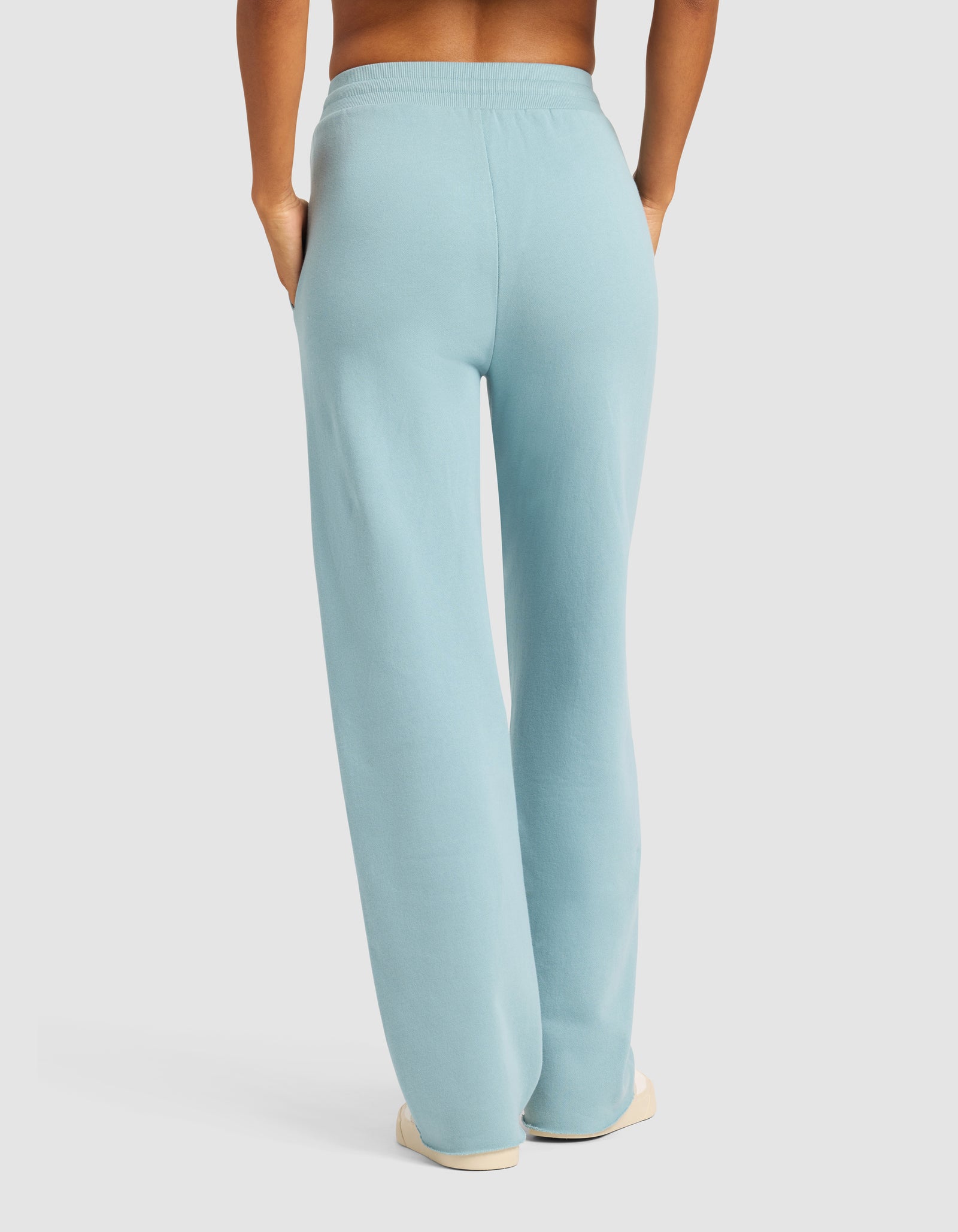 Model wearing Cozy Earth Chelsea Pant in light blue, paired with white shoes. Shown from the back with hands in pockets against a plain light background.