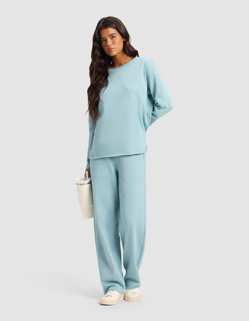 A woman with long dark hair wears the Cozy Earth Chelsea Pant, styled with a light blue long-sleeve top and beige sneakers. She holds a white tumbler and stands against a plain light gray background.