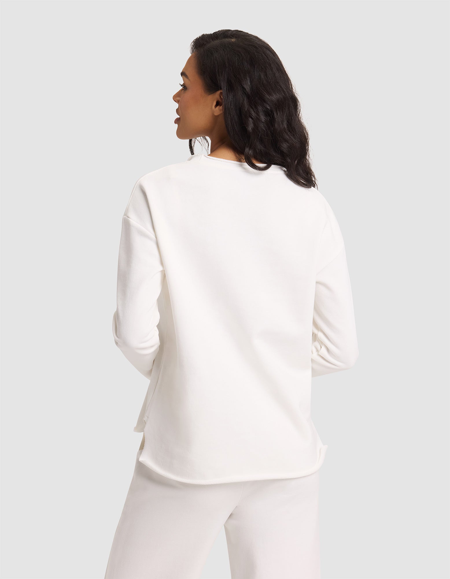 A woman with medium-length dark hair is seen from behind wearing Cozy Earth's Women's Chelsea Pullover in white, paired with white pants, standing against a plain light gray background. 