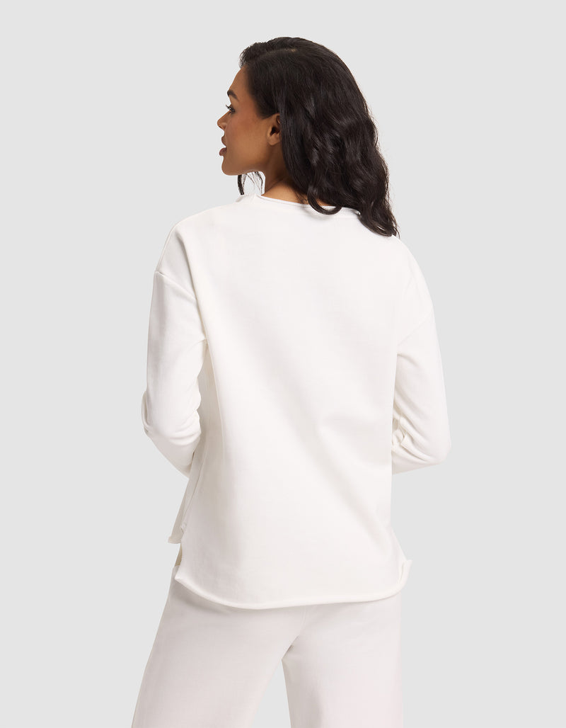 A woman with medium-length dark hair is seen from behind wearing Cozy Earth's Women's Chelsea Pullover in white, paired with white pants, standing against a plain light gray background.