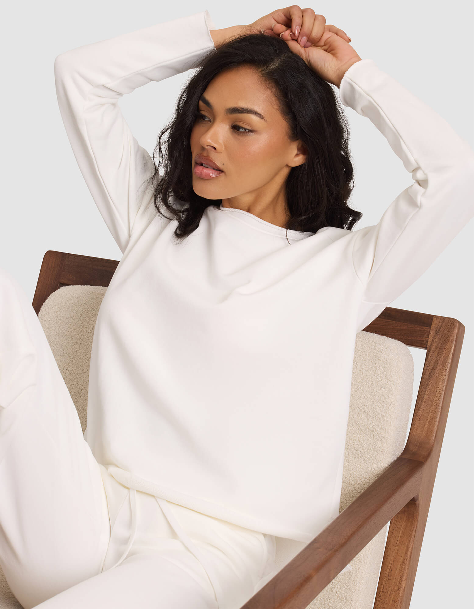 A woman with medium-length dark hair sits on a wooden chair, dressed in the Cozy Earth Women's Chelsea Pullover. With one arm raised and the other behind her head, she looks relaxed and gazes slightly to the side. 