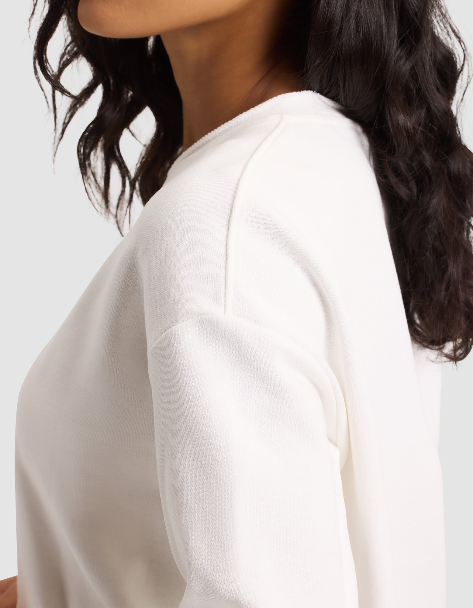 A woman with wavy dark hair wears the Cozy Earth Women's Chelsea Pullover, an off-white long-sleeve top, shown from the shoulders up and slightly turned to the side against a plain light background. 