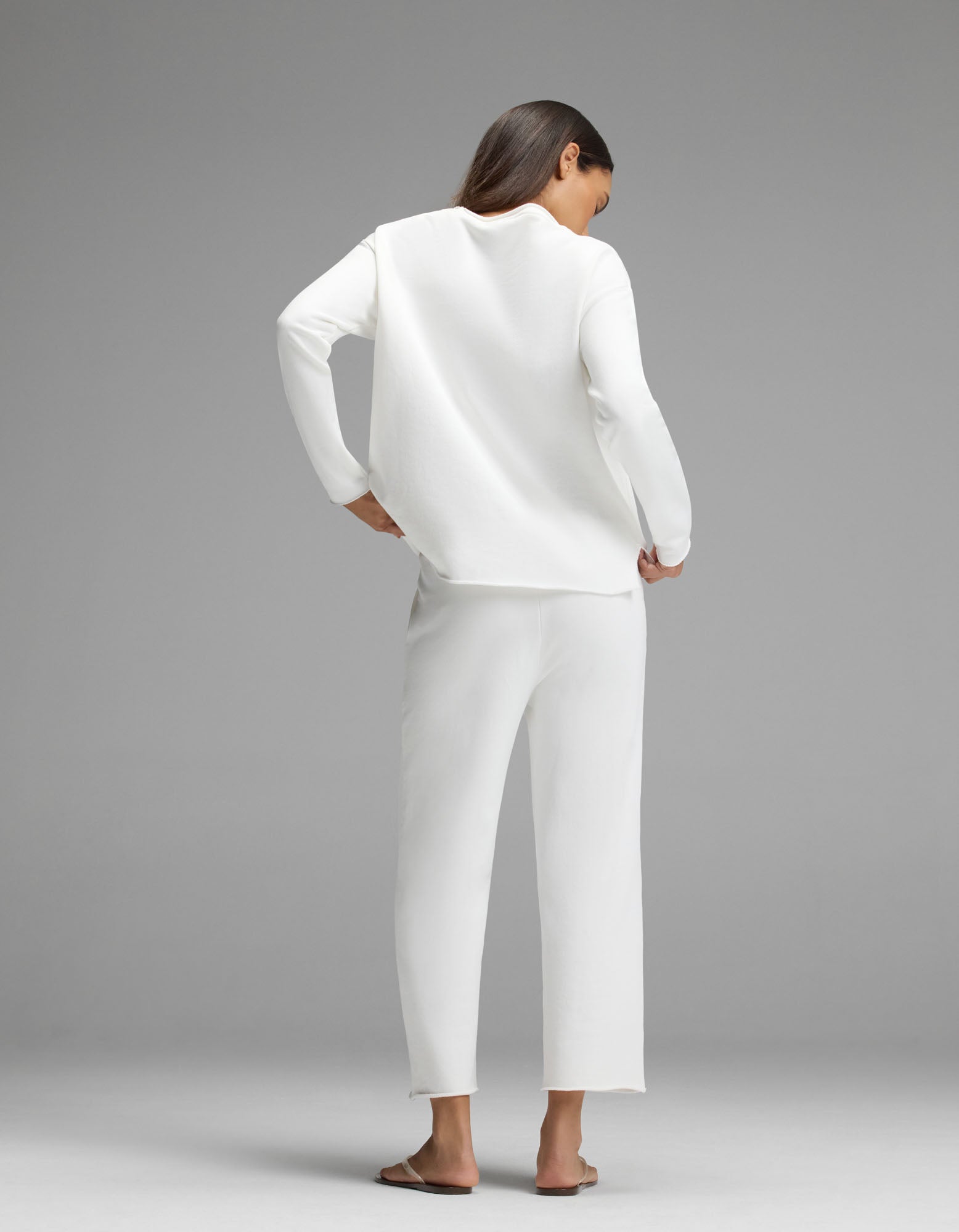 A woman stands with her back to the camera, barefoot, wearing the Cozy Earth Women’s Chelsea Cropped Set—an elegant, loose-fitting white long-sleeve top paired with matching white pants—against a plain gray background.
