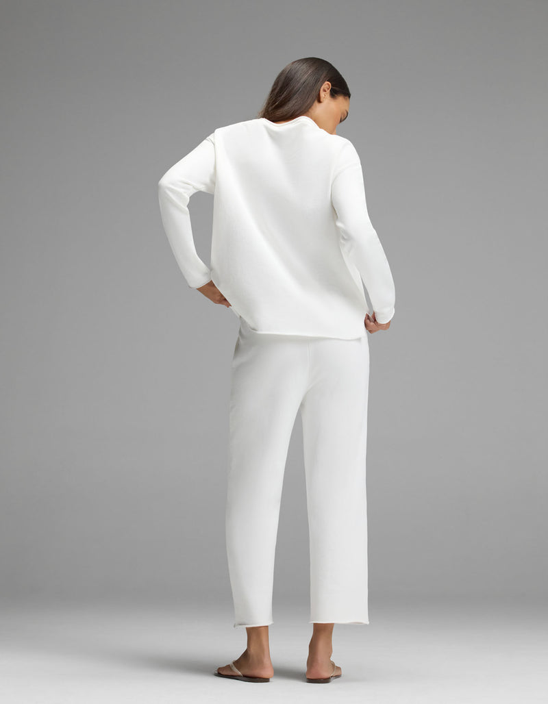 A woman stands with her back to the camera, barefoot, wearing the Cozy Earth Women’s Chelsea Cropped Set—an elegant, loose-fitting white long-sleeve top paired with matching white pants—against a plain gray background.