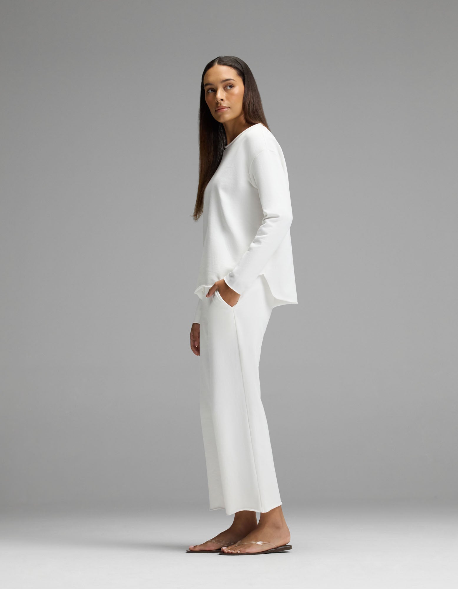 A woman stands sideways, looking forward, wearing the Cozy Earth Women’s Chelsea Cropped Set— a loose-fitting white long-sleeve top paired with matching wide-leg pants and sandals. The background is plain gray.