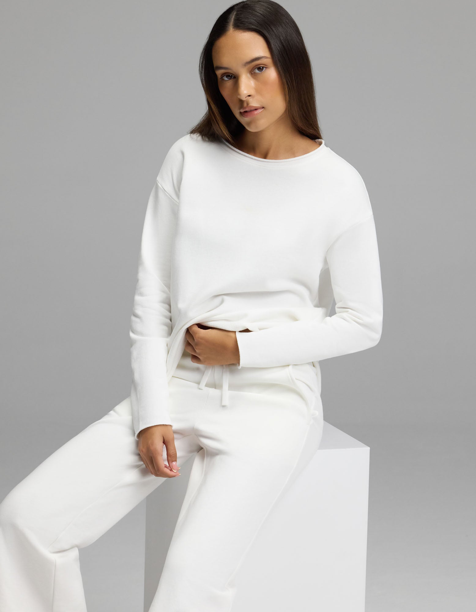 A woman with long, straight hair wears the Cozy Earth Women’s Chelsea Cropped Set—an all-white long-sleeve top and matching pants—as she sits on a white cube against a plain gray background, looking at the camera with a neutral expression.