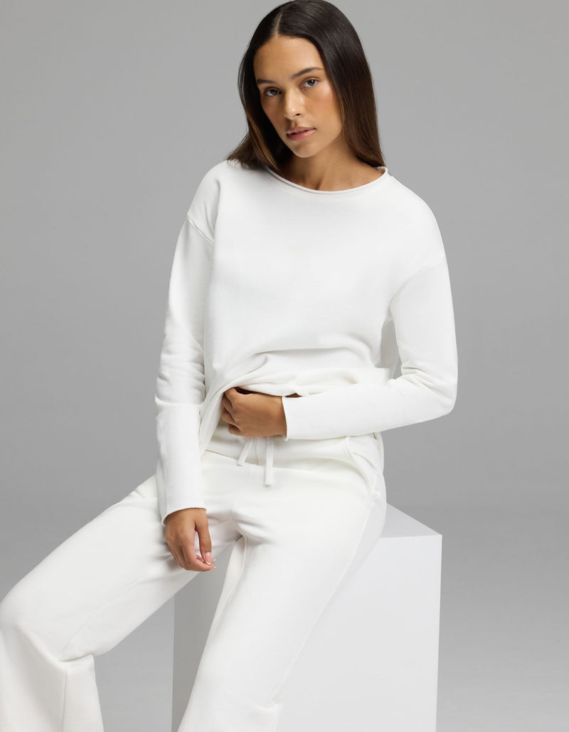 A woman with long, straight hair wears the Cozy Earth Women’s Chelsea Cropped Set—an all-white long-sleeve top and matching pants—as she sits on a white cube against a plain gray background, looking at the camera with a neutral expression.