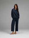 A woman stands in a relaxed pose, hands in her pockets, wearing the Cozy Earth Women’s Chelsea Cropped Set—a matching navy blue long-sleeve top and wide-leg pants—with brown sandals, against a plain gray background.|Color:Eclipse