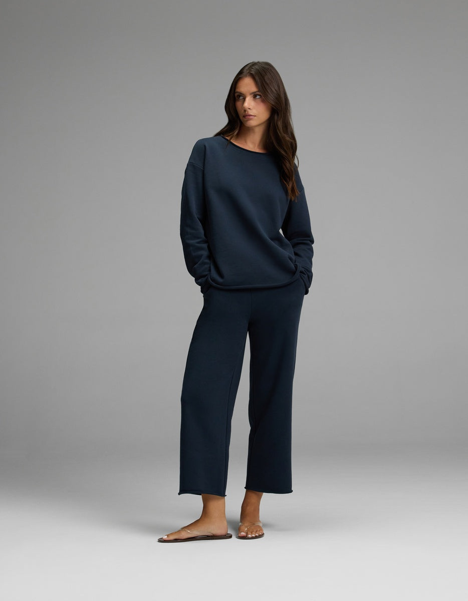 A woman stands in a relaxed pose, hands in her pockets, wearing the Cozy Earth Women’s Chelsea Cropped Set—a matching navy blue long-sleeve top and wide-leg pants—with brown sandals, against a plain gray background.|Color:Eclipse