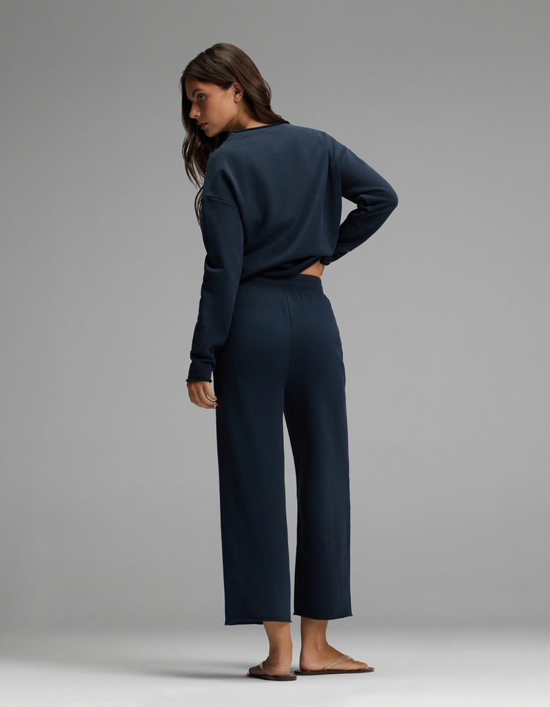 A woman with long brown hair stands barefoot on a light gray floor against a plain gray background, facing away and looking over her left shoulder. She is wearing the Cozy Earth Women’s Chelsea Cropped Set in dark blue, featuring a long-sleeve top and matching pants.