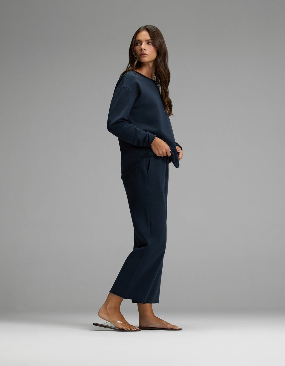 A woman stands sideways against a plain gray background, wearing the Women’s Chelsea Cropped Set in navy blue from Cozy Earth, paired with clear sandals.