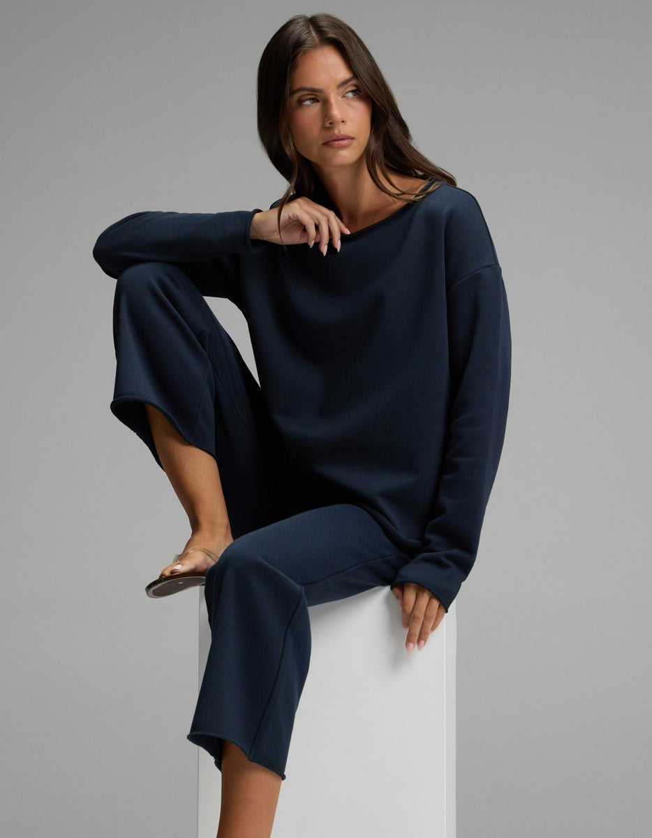 Wearing the Cozy Earth Women’s Chelsea Cropped Set, a woman sits on a white block with one knee raised and the other leg hanging down, gazing to the side against a plain gray background.