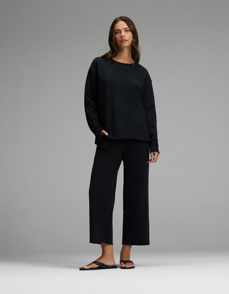 A woman stands against a plain background wearing the Cozy Earth Women’s Chelsea Cropped Set—featuring a loose black long-sleeve top and matching wide-leg pants—and black sandals, with one hand in her pocket.|Color:Jet Black