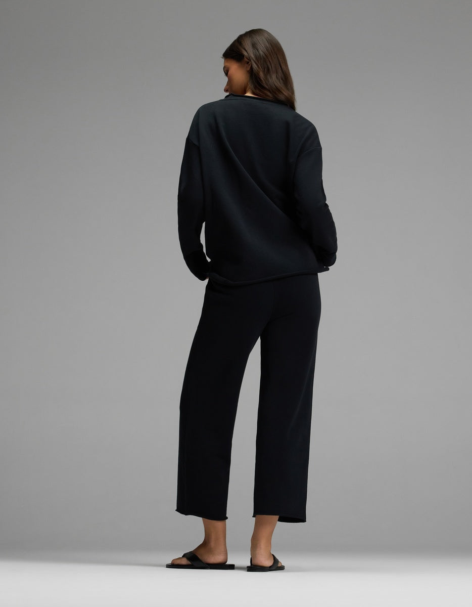 A person stands facing away, wearing the Cozy Earth Women’s Chelsea Cropped Set—a loose black long-sleeve top and matching wide-leg pants—with black sandals against a plain gray background.
