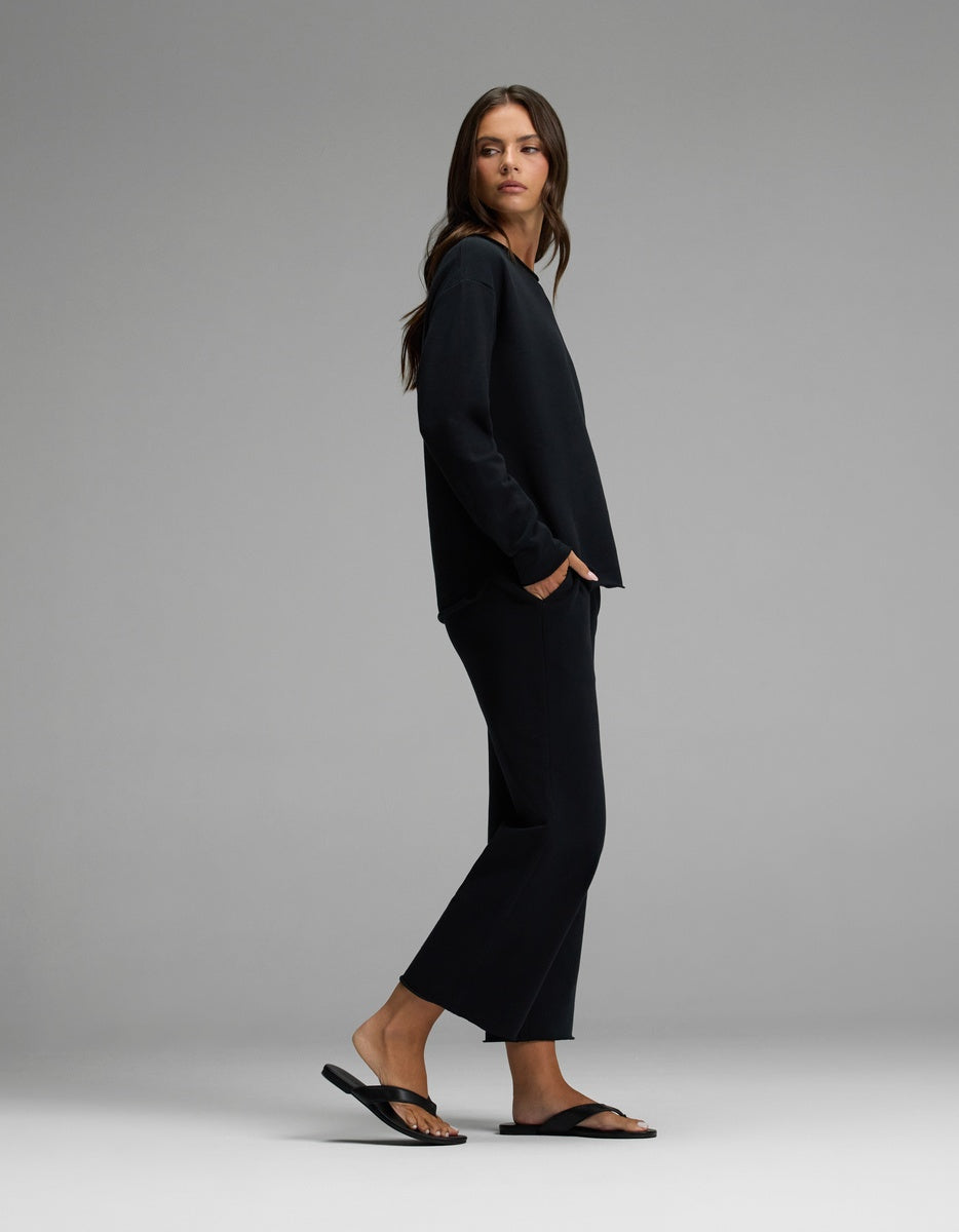 A woman stands sideways wearing the Cozy Earth Women’s Chelsea Cropped Set, consisting of a loose black long-sleeve top and matching black pants. She pairs the outfit with black sandals, has her hands in her pockets, and looks forward with a neutral expression against a plain gray background.