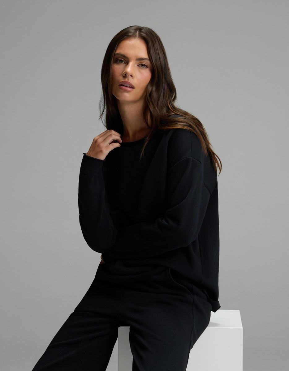 A woman with long brown hair wearing the Cozy Earth Women’s Chelsea Cropped Set in black sits on a white block, looking into the camera with a neutral expression against a plain gray background.