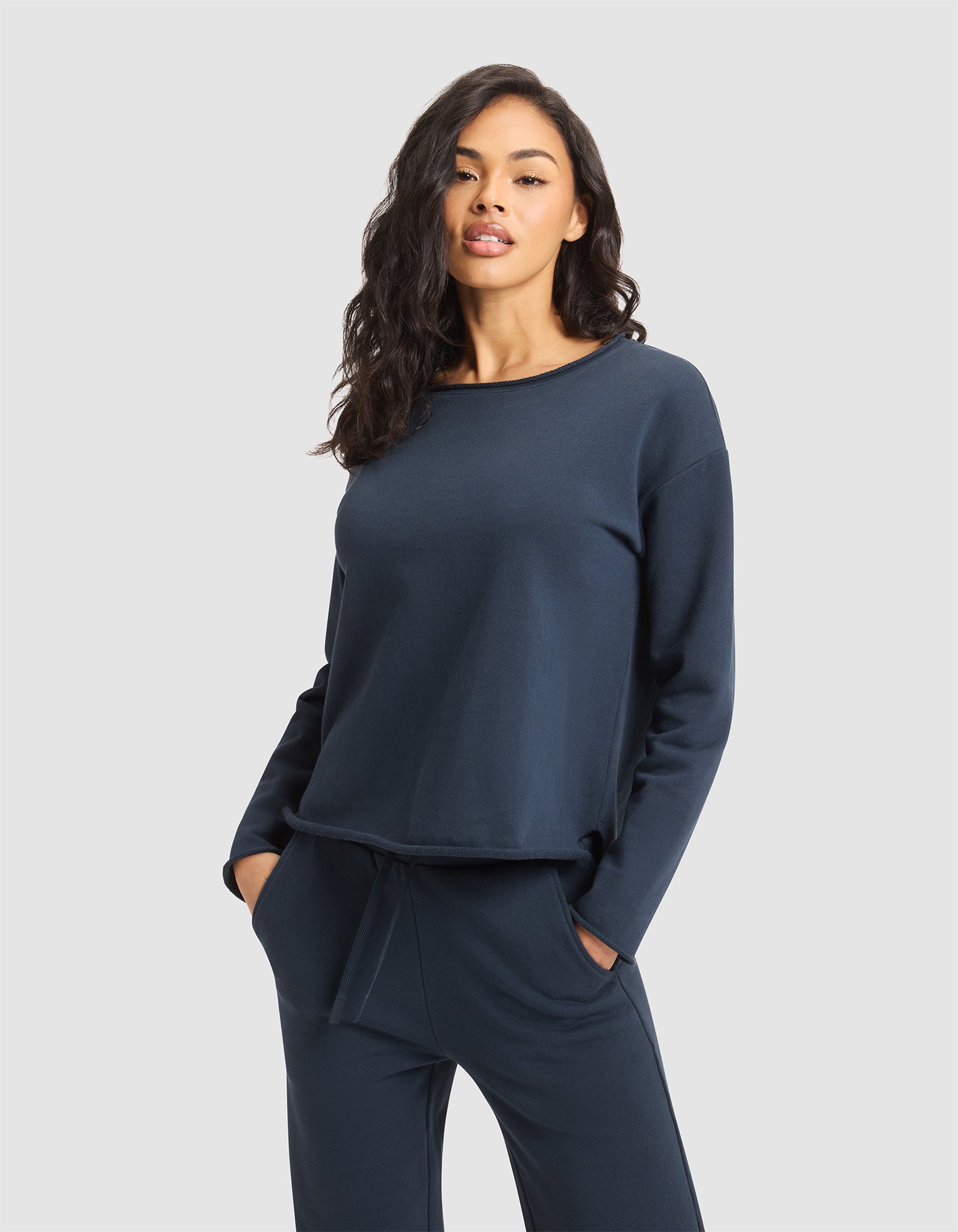 A woman with long, wavy dark hair wears the Cozy Earth Women's Chelsea Pullover and matching navy pants, standing with hands in her pockets against a plain light gray background.  
