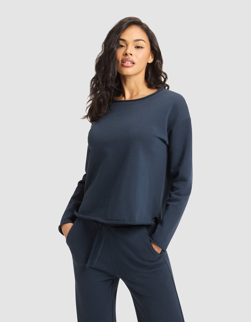 A woman with long, wavy dark hair wears the Cozy Earth Women's Chelsea Pullover and matching navy pants, standing with hands in her pockets against a plain light gray background.
