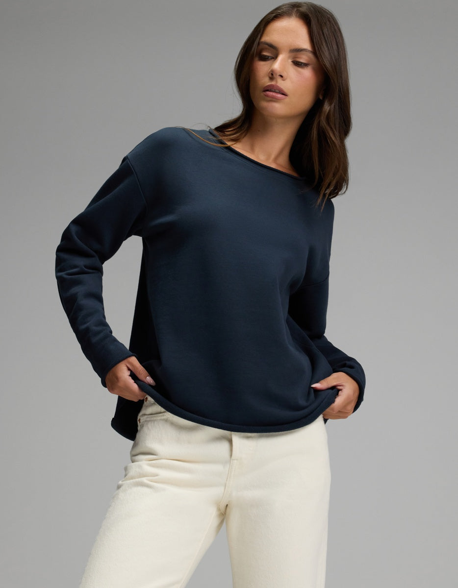 A woman wearing the Cozy Earth Women’s Chelsea Pullover in Eclipse and off-white pants stands against a plain gray background, looking to her left with her hands in her pockets.|Color:Eclipse