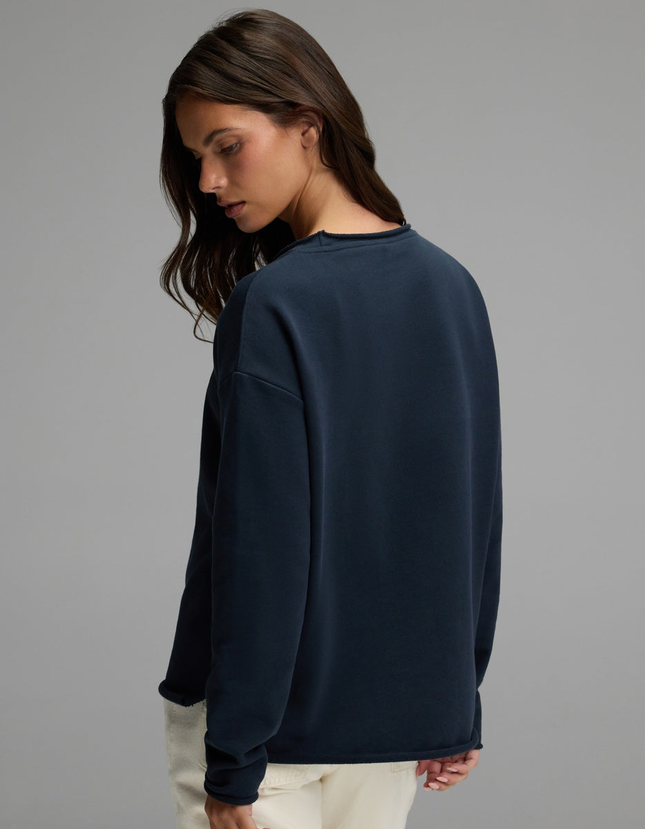 A woman with long brown hair stands against a plain gray background, looking over her shoulder while wearing the Cozy Earth Women’s Chelsea Pullover in dark blue and light pants.