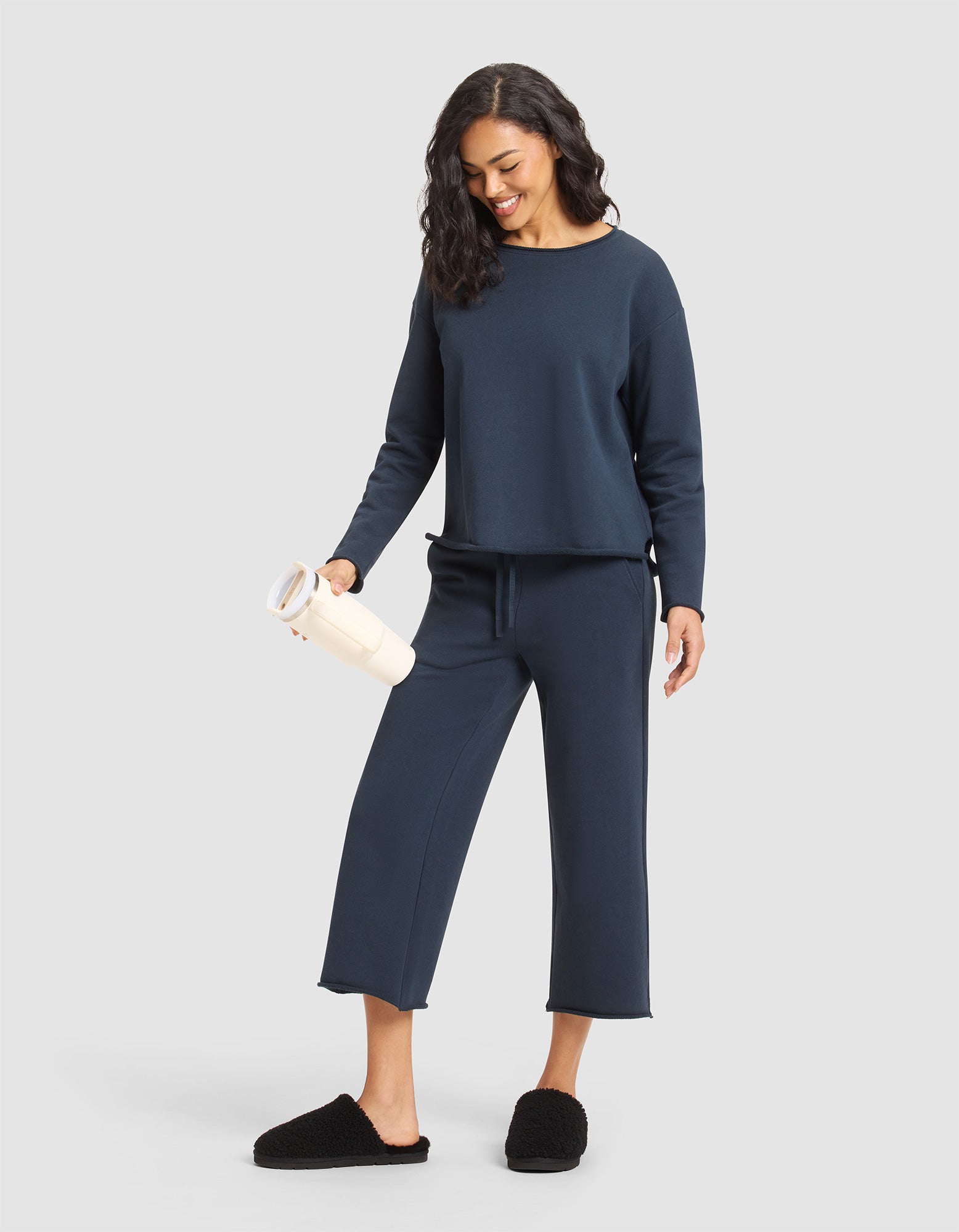 A woman wears the Cozy Earth Women's Chelsea Pullover in navy with matching wide-leg pants, paired with black slippers, and smiles while holding a light tumbler against a plain light gray background. 