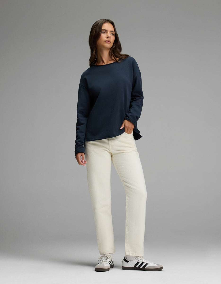 A woman stands against a gray background, wearing Cozy Earth’s Women’s Chelsea Pullover in navy blue, paired with off-white pants and white adidas sneakers with black stripes. She has straight brown hair and a neutral expression, with one hand in her pocket.