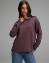 A woman wearing the Cozy Earth Women’s Chelsea Pullover in Fig and blue jeans stands against a plain gray background, looking slightly to the side with one hand near her neck.|Color:Fig