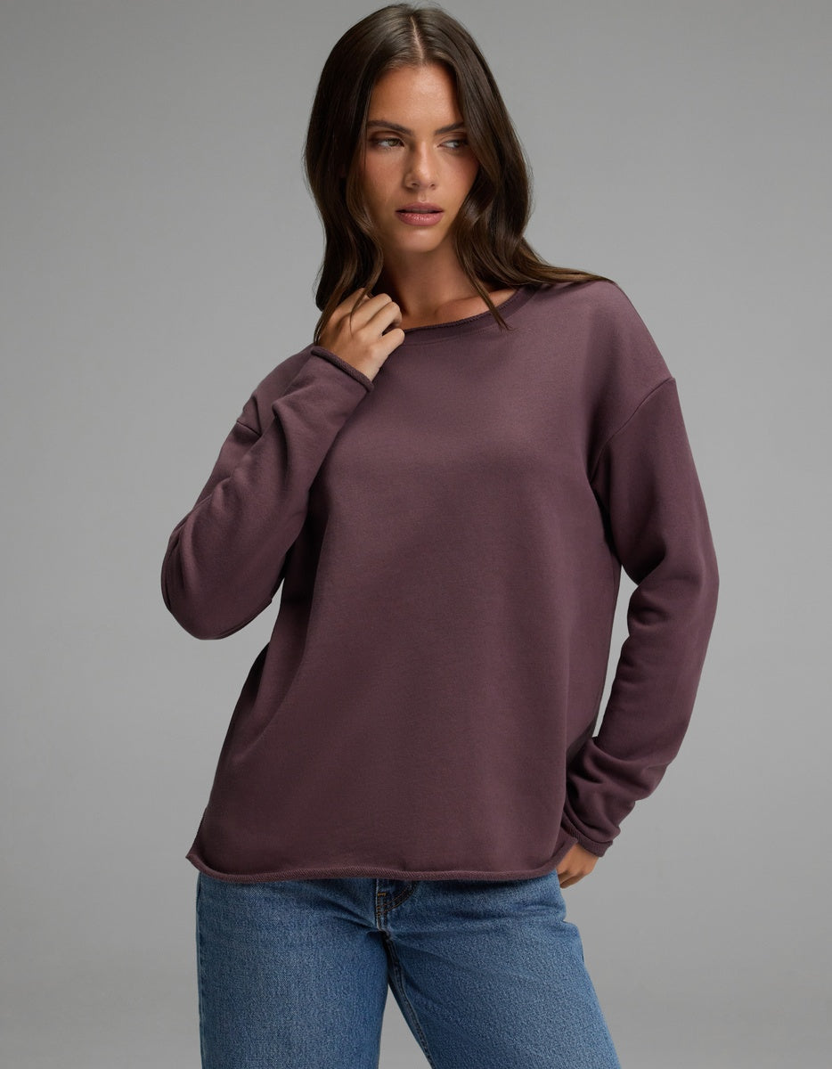 A woman wearing the Cozy Earth Women’s Chelsea Pullover in Fig and blue jeans stands against a plain gray background, looking slightly to the side with one hand near her neck.