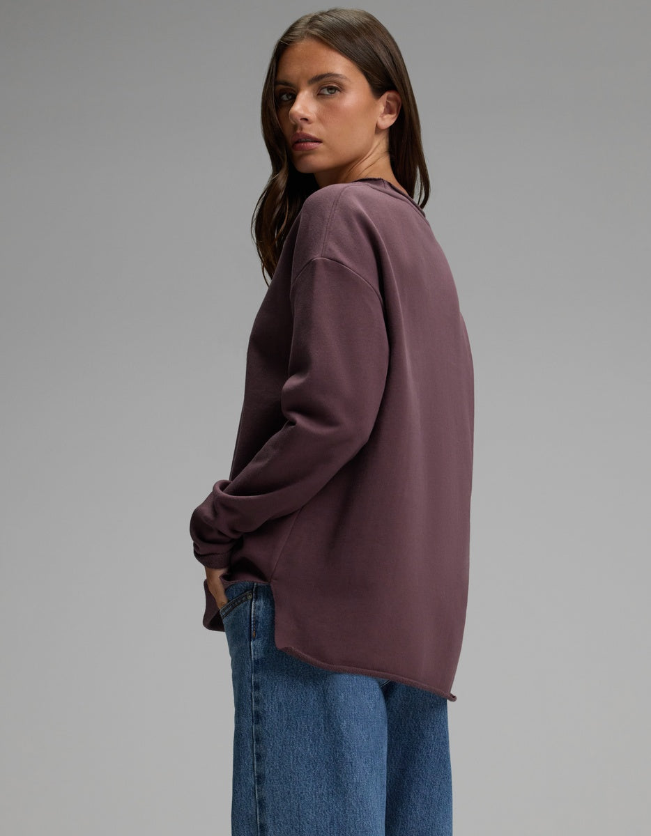 A person with long brown hair stands sideways and looks over their shoulder against a plain gray background, wearing the Cozy Earth Women’s Chelsea Pullover in purple paired with blue jeans.