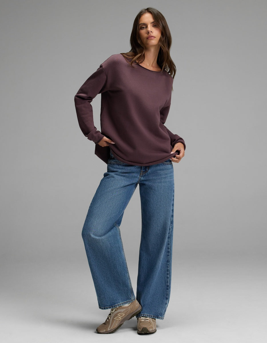 A woman with long brown hair and a relaxed expression poses in a simple studio, wearing the Cozy Earth Women’s Chelsea Pullover—a loose, dark purple sweatshirt—paired with wide-leg blue jeans and tan sneakers.